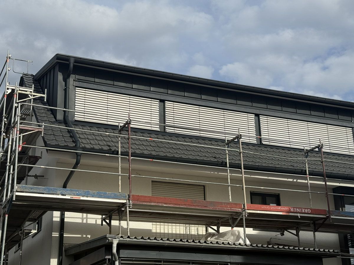 Ein weißes Gebäude, dessen Fassade renoviert wird, mit Gerüst und einem großen Dachgaubenfenster mit horizontalen Jalousien.