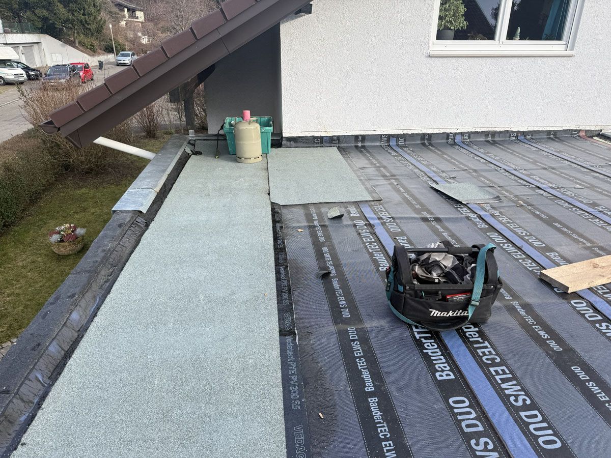 Ein im Bau befindliches Flachdach mit grauen Dachmembranen, Dämmplatten und einer Werkzeugtasche auf der Oberfläche.