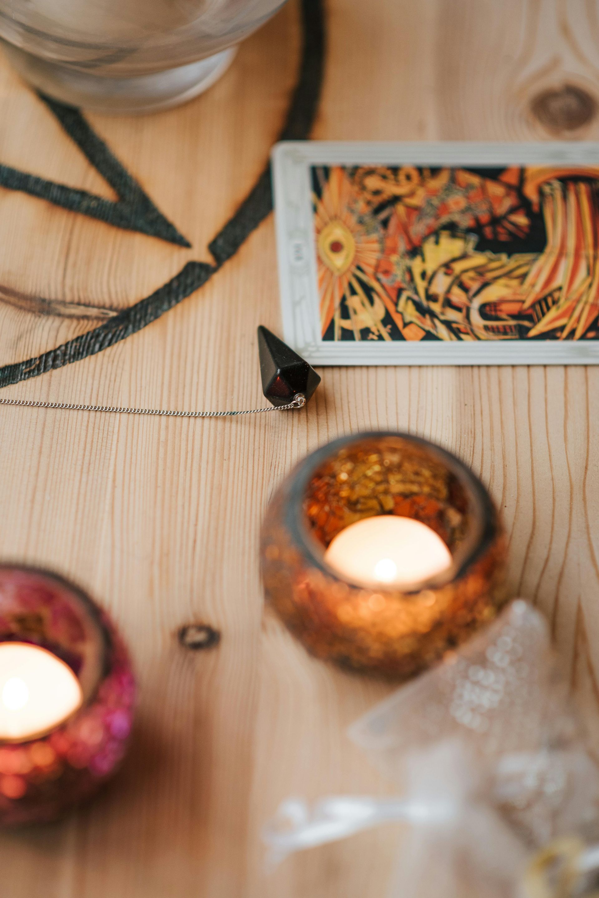 Velas, carta de tarot, péndulo de cristal y elementos decorativos sobre una superficie de madera.