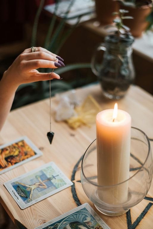 Persona sosteniendo un péndulo sobre cartas del tarot y una vela encendida sobre una mesa de madera.