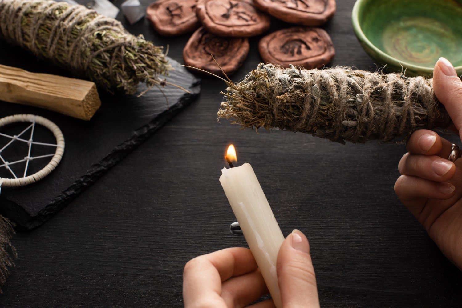 Manos encendiendo un manojo de salvia con una vela sobre una mesa oscura, rodeada de elementos espirituales.
