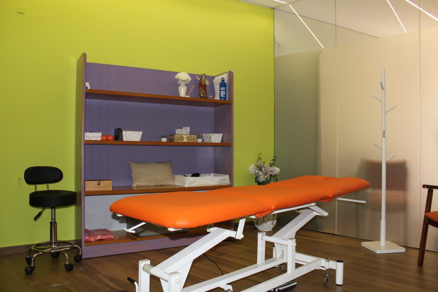 Mesa de masaje naranja en una sala médica con estantes morados y pared verde.