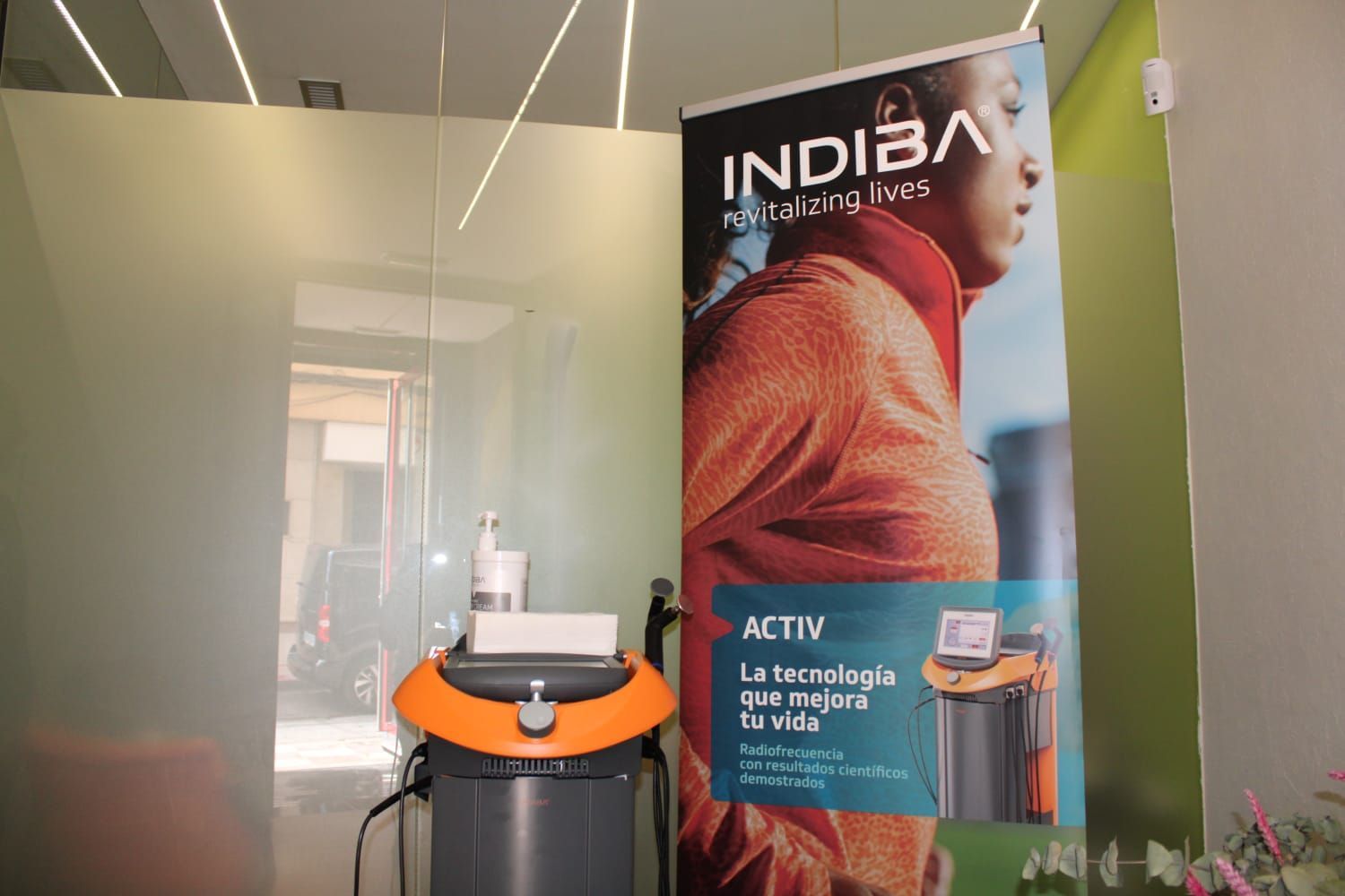 Máquina INDIB Activ junto a pancarta promocional y mampara de cristal; ambientación interior con colores neutros.