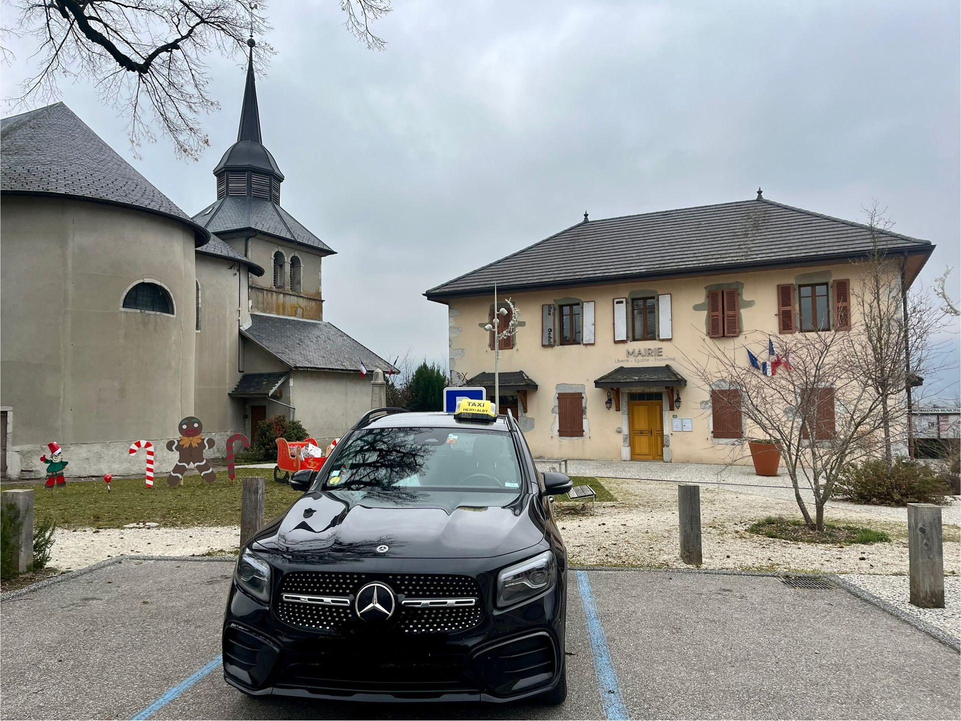 Un SUV Mercedes noir est garé devant une église et un bâtiment orné de drapeaux ; temps nuageux.