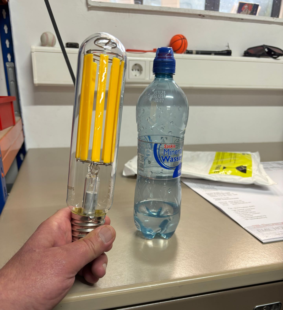 Eine Hand hält eine gelbe LED-Lampe in einem Raum; im Hintergrund sind eine Wasserflasche und Utensilien zu sehen.