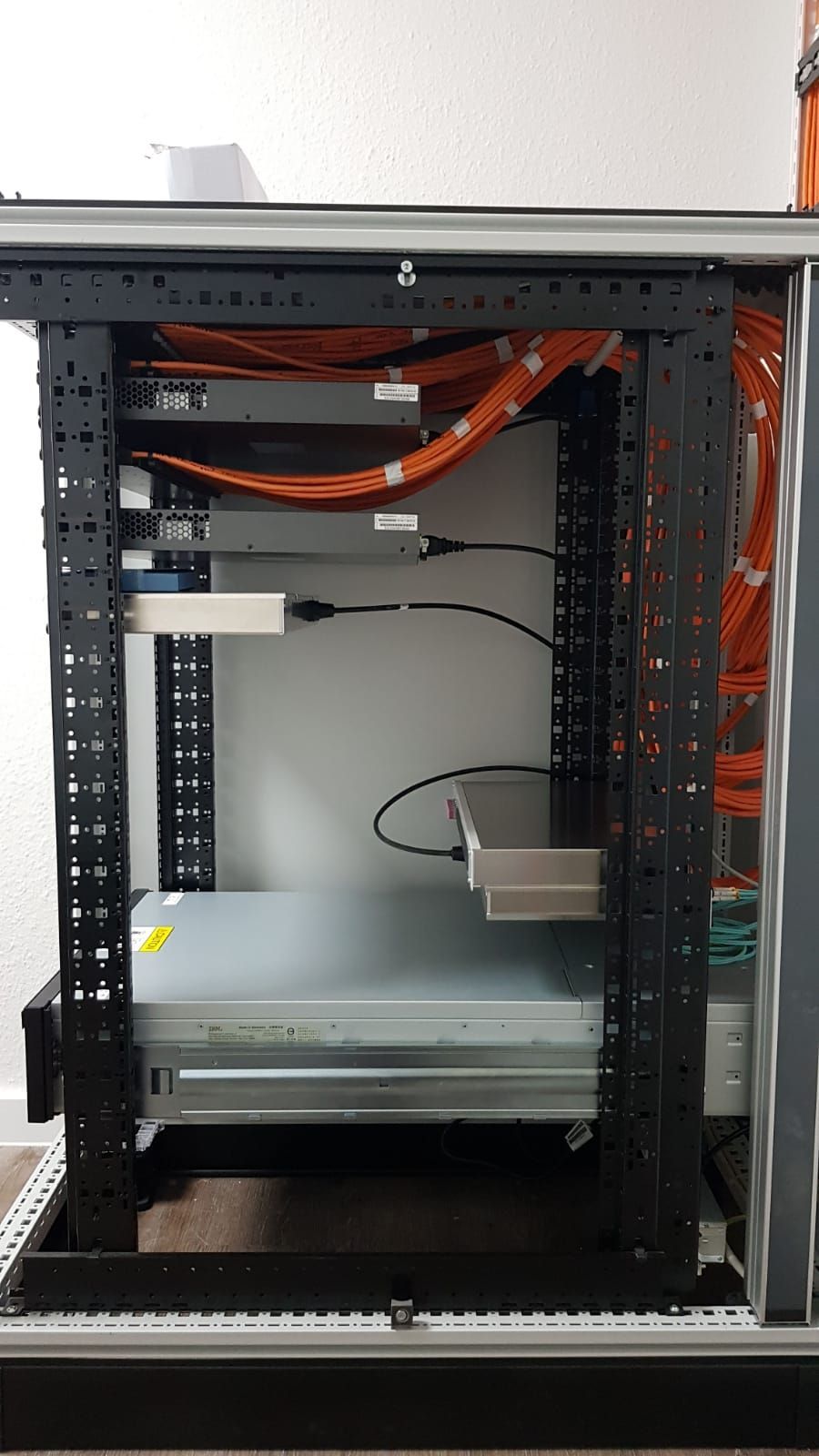 Offenes Netzwerk-Rack mit orangefarbenen Kabeln und eingebauten Geräten in einem grauen Schrank.
