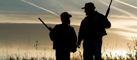 Dos cazadores recortados contra el cielo al atardecer en un campo de hierba, sosteniendo rifles