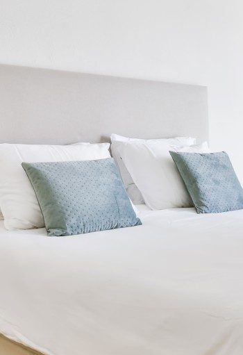Lit avec draps blancs, tête de lit grise et coussins décoratifs bleus.