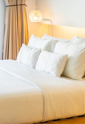 Lit blanc avec de nombreux oreillers et une tête de lit beige, près d'un rideau.