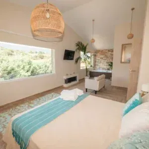 Dormitorio luminoso con gran ventanal, cama con manta verde azulado, lámparas tejidas y una pequeña chimenea.