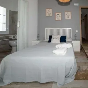 Dormitorio con cama, mesitas de noche y cuadros en una pared gris. Baño visible tras una puerta.