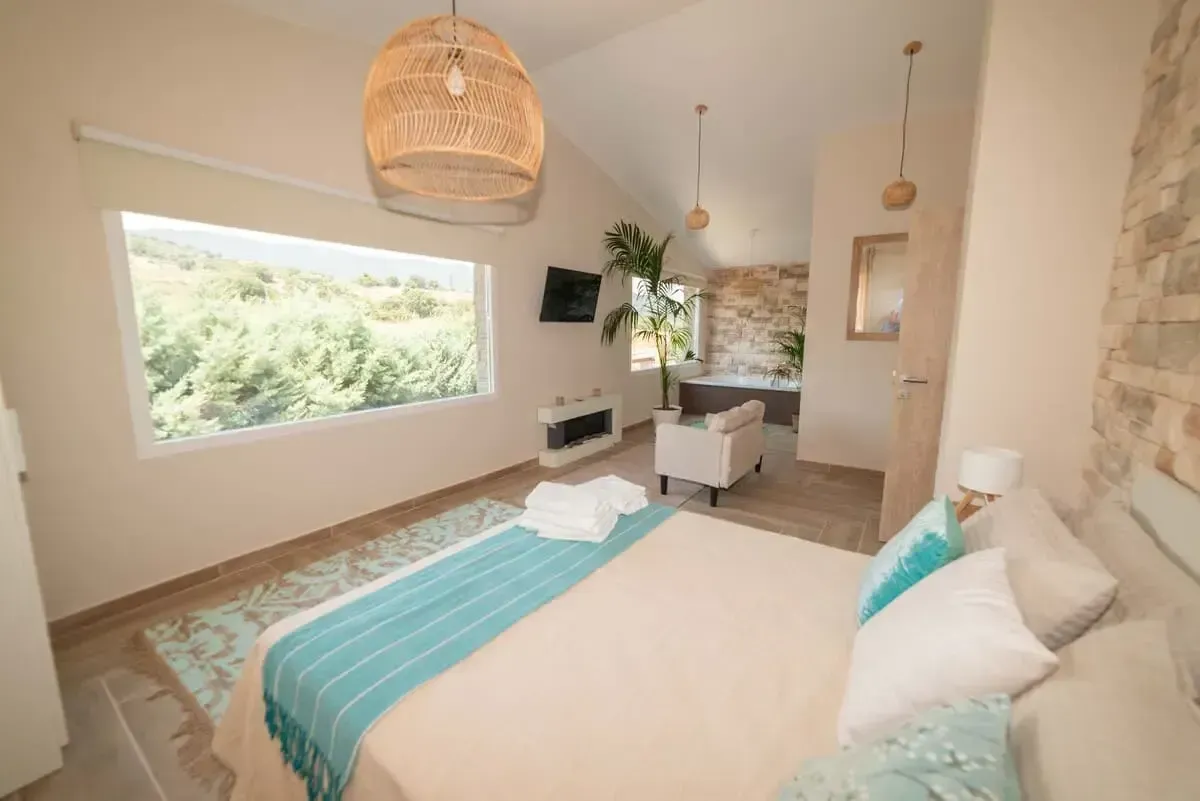 Dormitorio con cama, manta azul, alfombra, gran ventanal con vista a los árboles y lámpara colgante de mimbre.