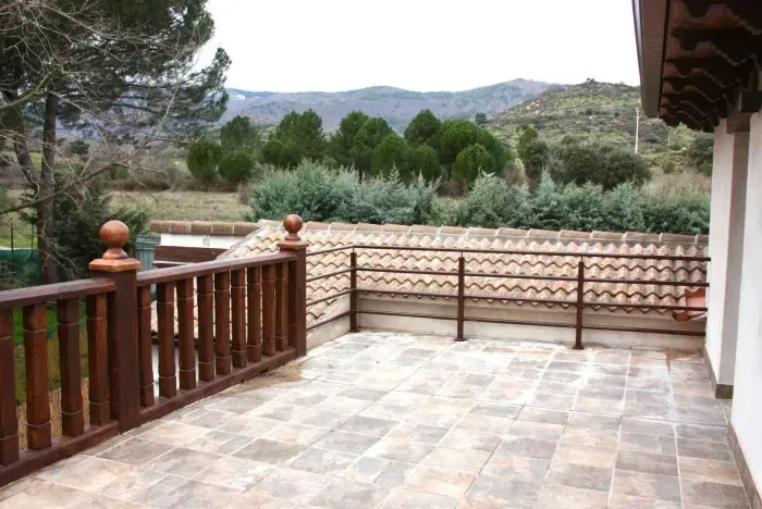 Patio con barandilla marrón y suelo de baldosas, con vista a árboles y montañas.