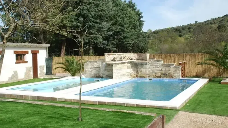 Piscina al aire libre de dos niveles con cascada de piedra, rodeada de césped verde y árboles.