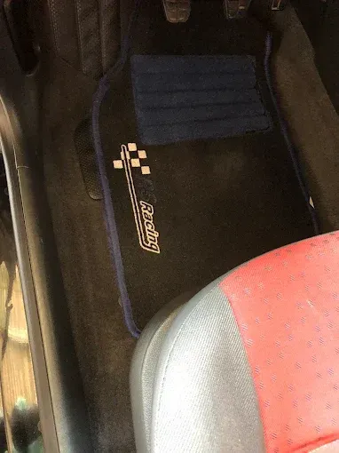 Alfombrilla negra para coche con ribete azul junto a un cojín de asiento rojo y gris.