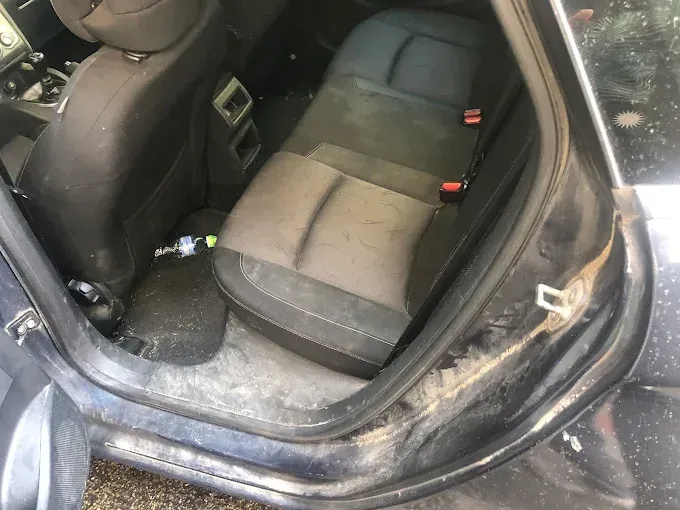 Asiento trasero del coche sucio con tapicería gris y suciedad visible en el suelo y el umbral de la puerta.