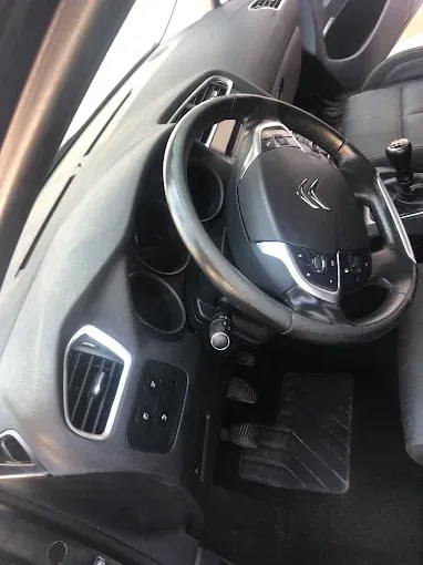 Interior del coche con volante de cuero negro, controles del salpicadero y alfombrilla visibles desde el lado del conductor.