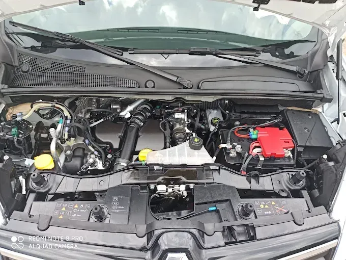 Capó abierto que deja ver el compartimento del motor con componentes negros y una tapa de batería roja a la derecha.