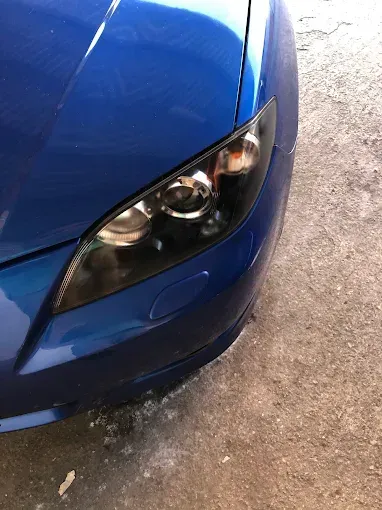 Primer plano del faro delantero y el parachoques de un coche azul sobre pavimento mojado.