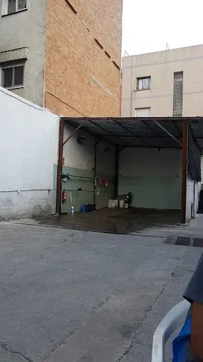 Pequeño garaje abierto situado en un patio entre edificios, con techo de metal y objetos dispersos en su interior.