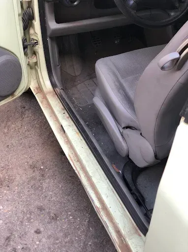 Interior del coche con la puerta abierta, mostrando un asiento gris y el umbral de la puerta desgastado junto al pavimento.