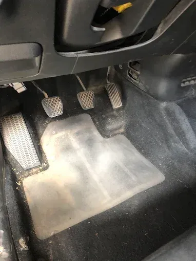 Piso del auto con pedales y una alfombrilla de goma polvorienta en el lado del conductor