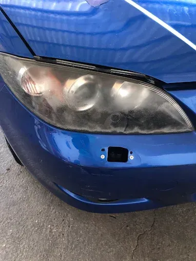 Esquina delantera de un coche azul con un faro dañado y sin la cubierta del parachoques.