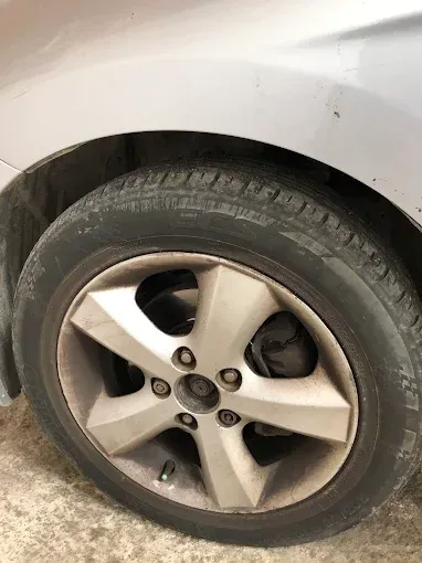 Primer plano de la rueda delantera y el neumático de un coche blanco, donde se aprecia una llanta de aleación sucia.