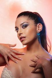 Mujer con cabello oscuro recogido en una cola alta, con sombra de ojos azul, tocando su pecho, con un fondo rosa.