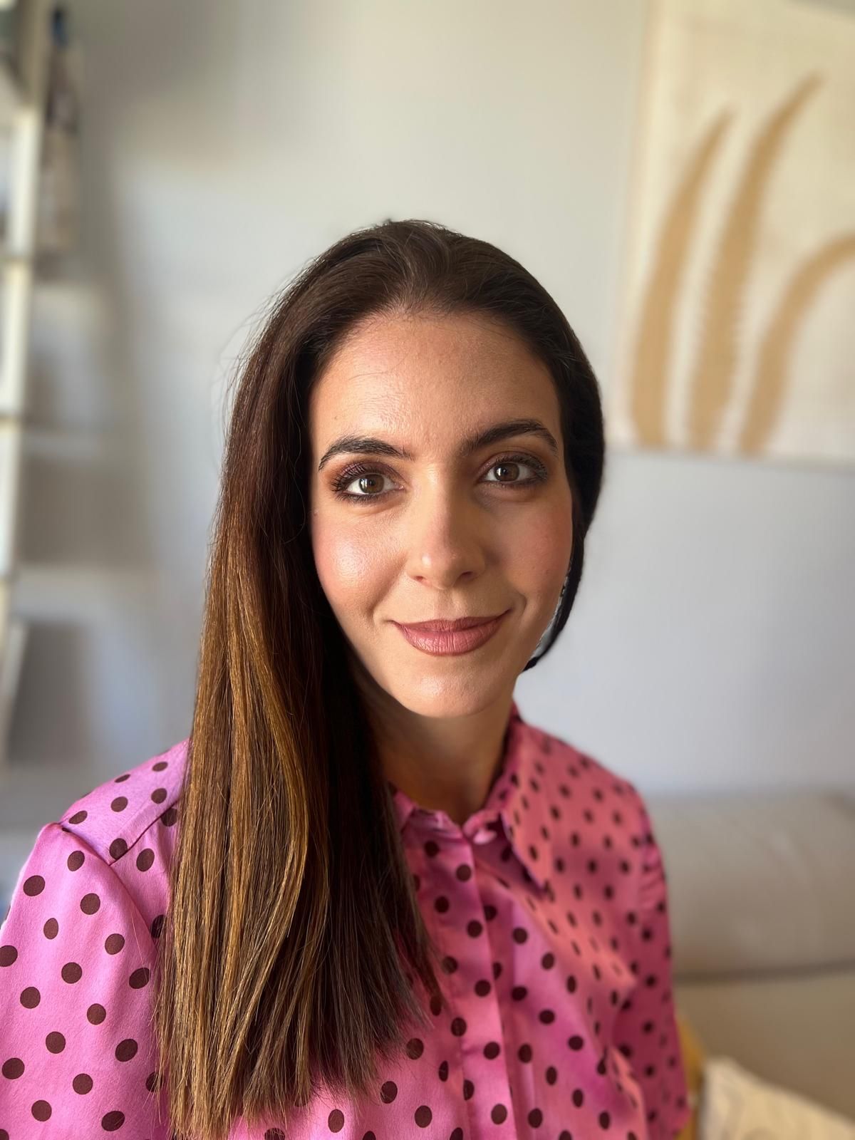 Mujer con cabello largo y castaño, vestida con una camisa de lunares rosas, sonriendo en una habitación.