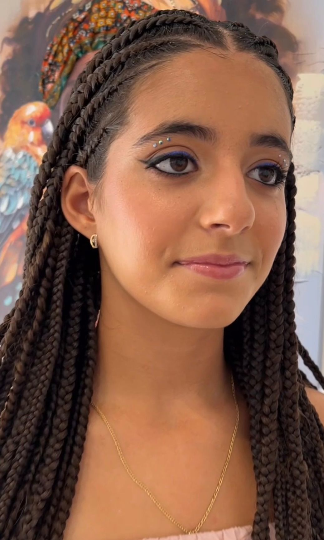 Mujer joven con cabello trenzado, delineador de ojos azul y joyas de oro, sonriendo.