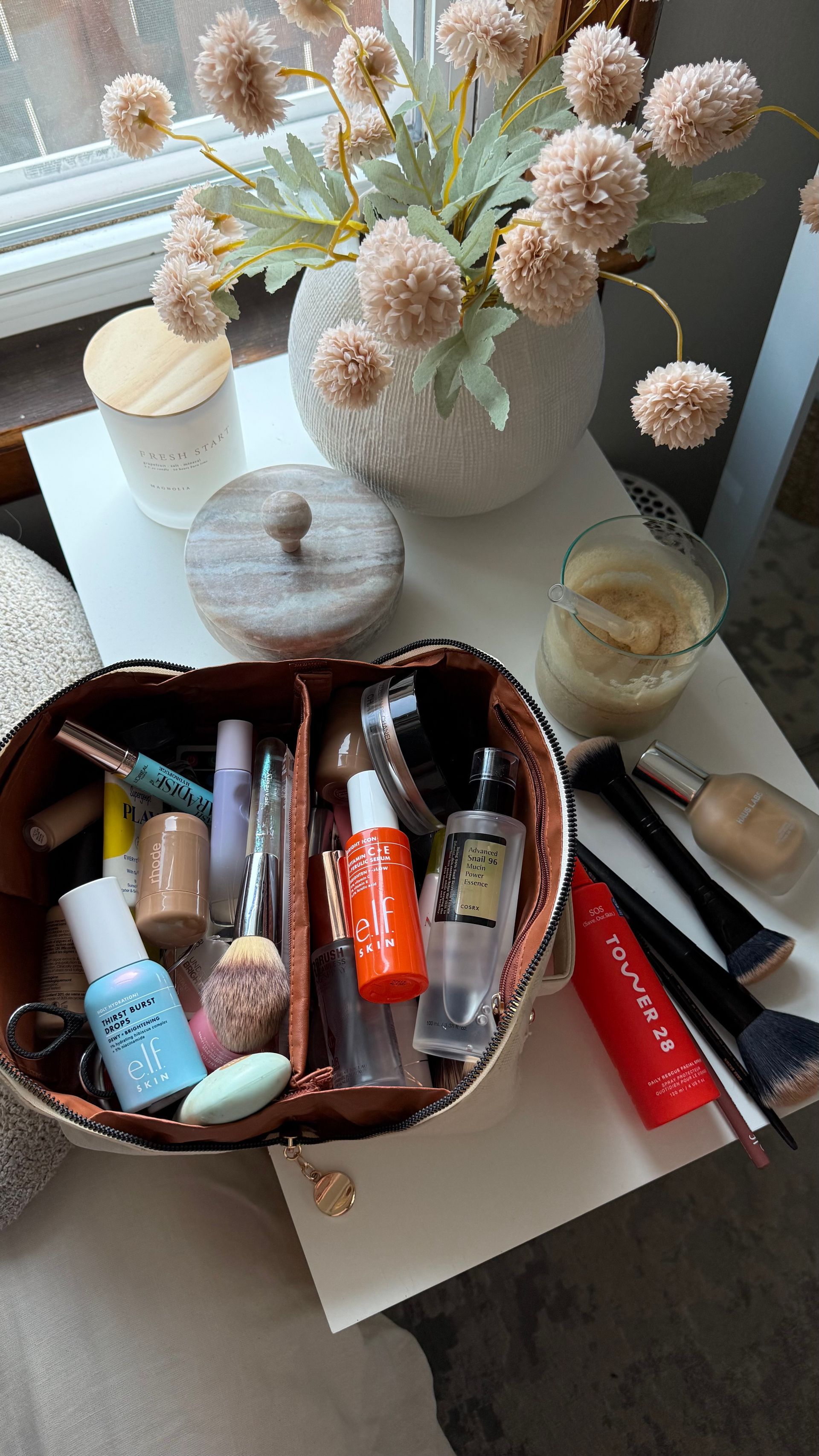 Bolsa de maquillaje con cosméticos y pinceles sobre una mesa blanca junto a un jarrón de flores.