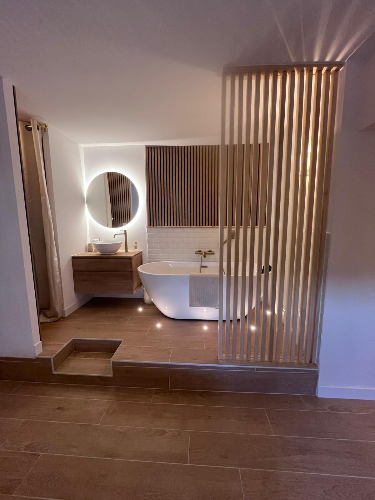 Salle de bains moderne avec baignoire îlot, miroir rond rétroéclairé, meuble-lavabo en bois et cloison verticale à lattes.