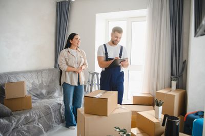 Zwei Personen packen in einem hellen Wohnzimmer Umzugskartons aus. Ein Sofa ist in Schutzfolie eingewickelt, die Vorhänge sind geöffnet.