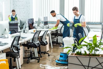 Reinigungskräfte wischen in einem überfluteten Büro den Boden. Um sie herum stehen Schreibtische, Monitore und Stühle