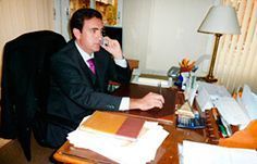 Un hombre con traje y corbata está sentado en un escritorio hablando por un teléfono celular.