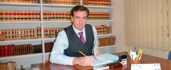 Un hombre con traje y corbata está sentado en un escritorio frente a una estantería.