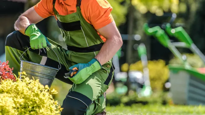 Jardinero con camisa naranja y delantal verde plantando flores en un jardín soleado.
