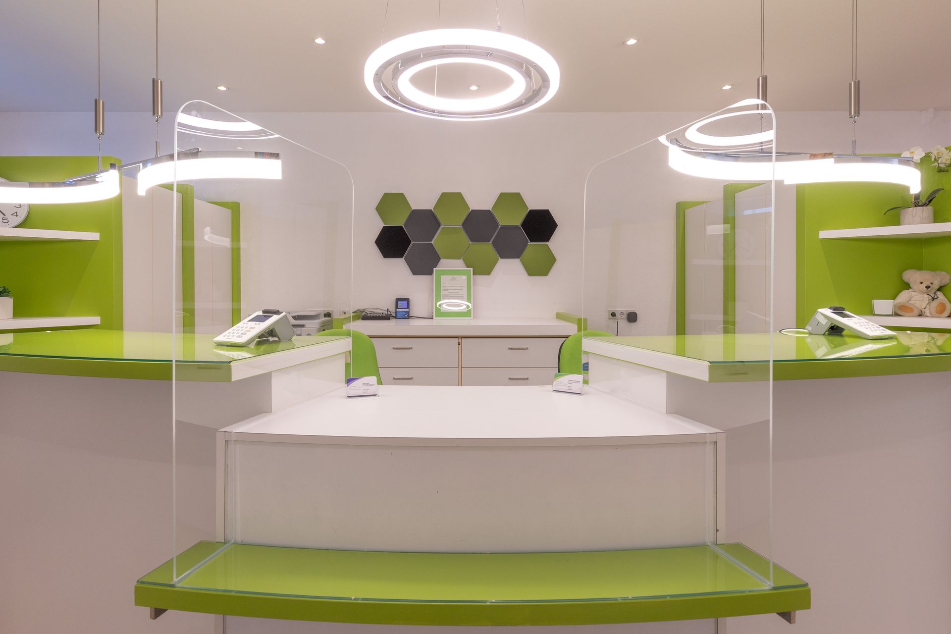 Moderner Empfangstresen in Weiß und Grün mit Schutzbarrieren, sechseckiger Wandkunst und Deckenbeleuchtung.