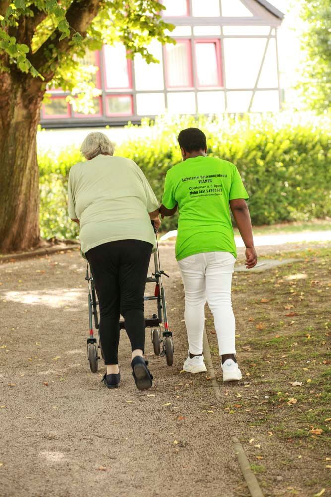 Eine ältere Person benutzt einen Rollator und wird dabei von einer Person in einem grünen Hemd auf einem Kiesweg im Freien unterstützt.