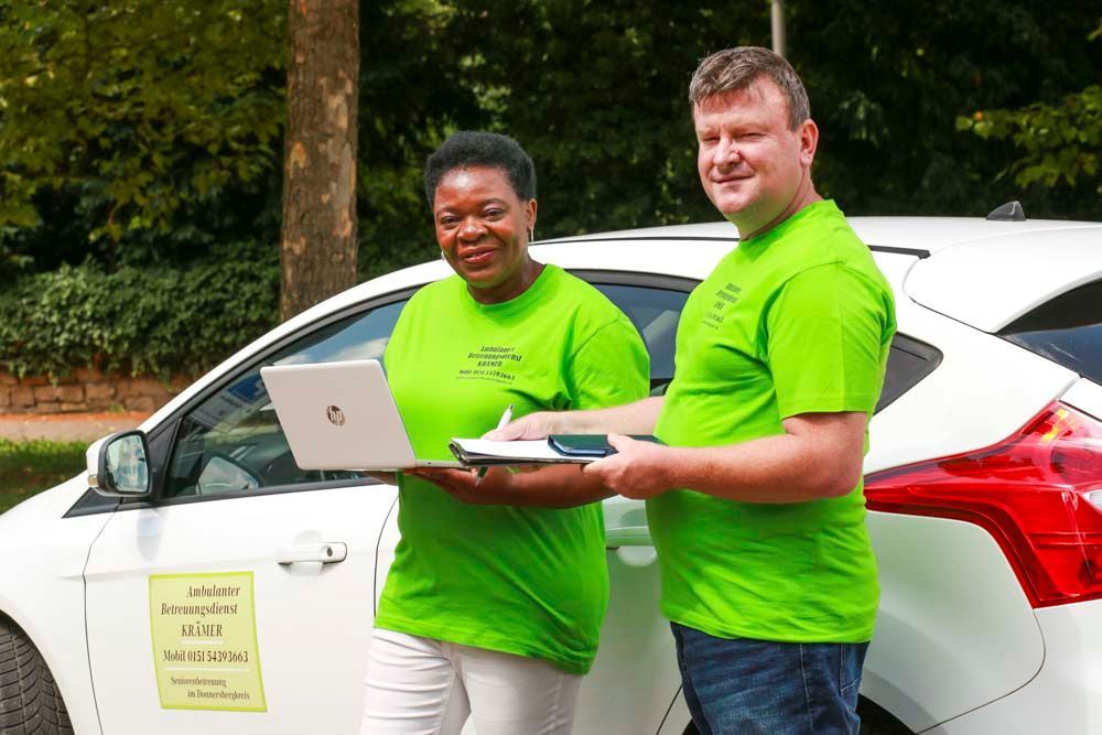 Zwei Personen in grünen Hemden neben einem weißen Auto, eine mit einem Laptop, die andere mit einem Notizblock.