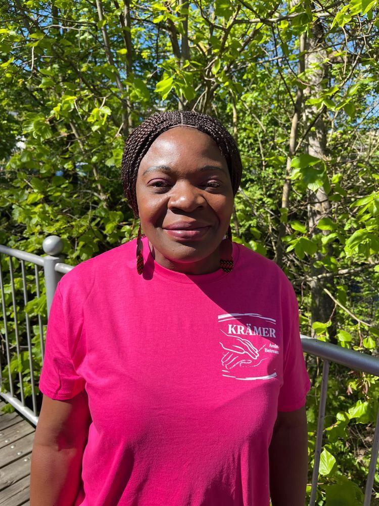 Frau in einem rosa T-Shirt steht im Freien an einem Metallgeländer und lächelt ins Sonnenlicht.