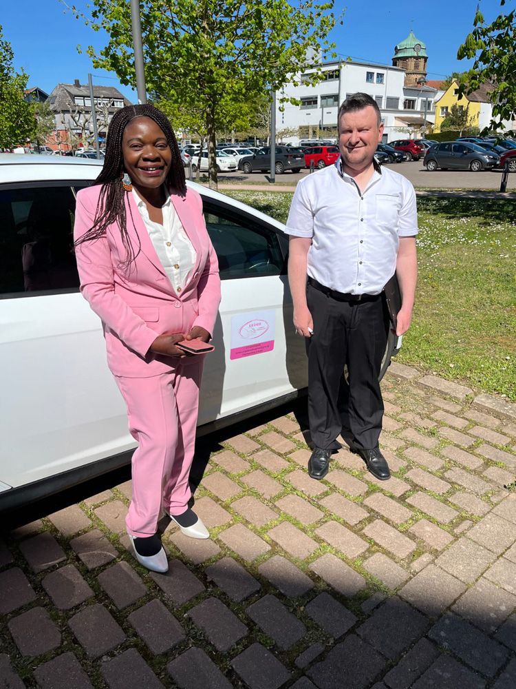 Zwei Personen stehen neben einem weißen Auto auf einem sonnigen Parkplatz; im Hintergrund sind Gebäude und Bäume zu sehen.