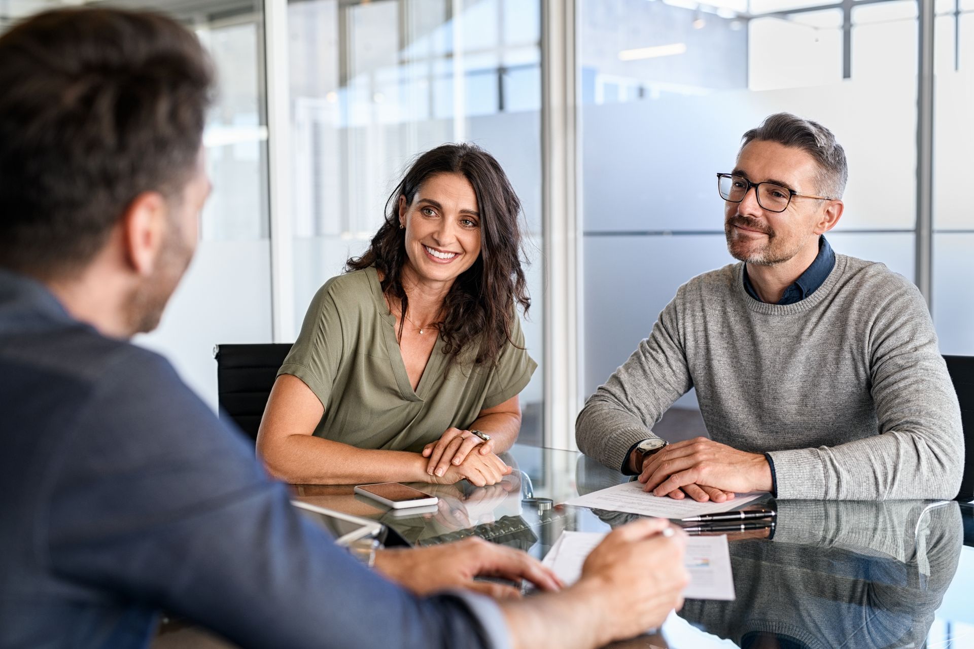 Trois personnes sont assises à une table, deux sourient, une parle, dans un bureau moderne aux parois de verre.