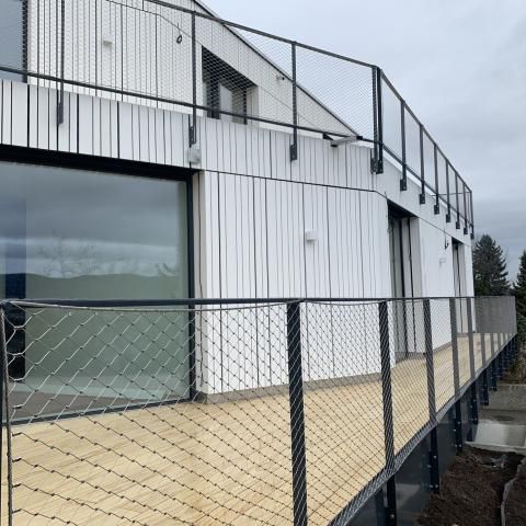 Modernes Gebäude mit Holzterrasse und schwarzem Metallgeländer mit Drahtgeflecht.