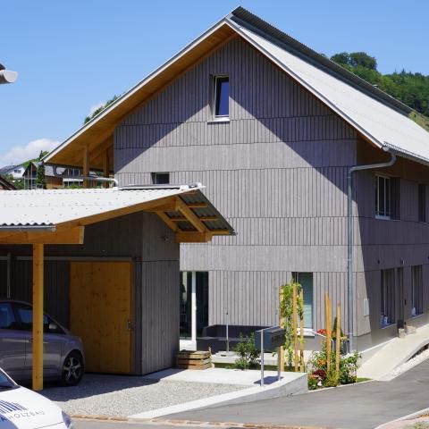 Graues Gebäude mit Carport. Ein Auto steht unter dem Carport. Im Hintergrund strahlend blauer Himmel.