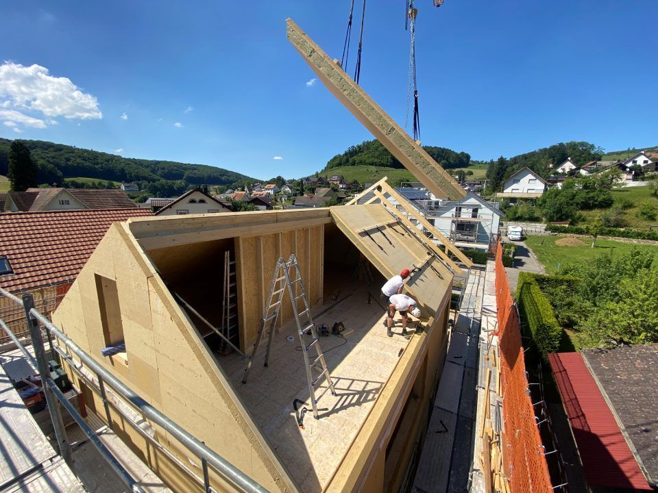 Baustelle: Ein Kran hebt einen Holzbalken auf ein Gebäude; Arbeiter sind im Hintergrund zu sehen; blauer Himmel, grüne Hügel.