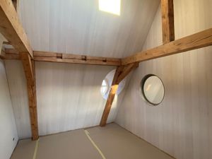 Zimmerinterieur mit freiliegenden Holzbalken, weissen Wänden und runden Fenstern.