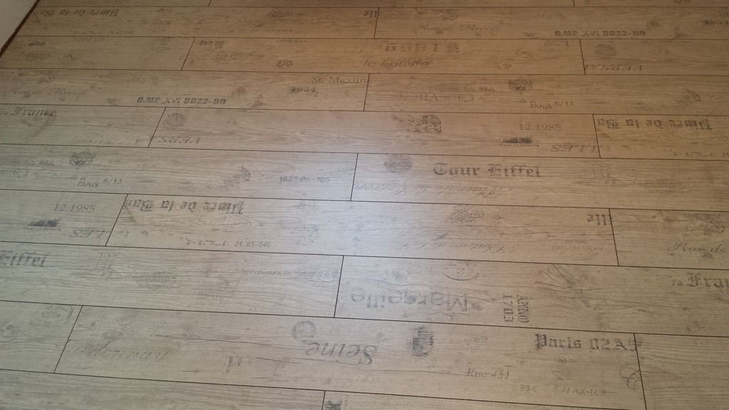 Hellbrauner Bodenbelag in Holzoptik mit verblasstem Text und gestrichelten Linien, wahrscheinlich zum Zuschneiden oder Verlegen.