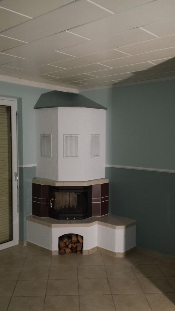 Weißer und kastanienbrauner Kamin in einem Raum mit hellblaugrünen Wänden, Brennholz im Sockel und einer Tür auf der linken Seite.